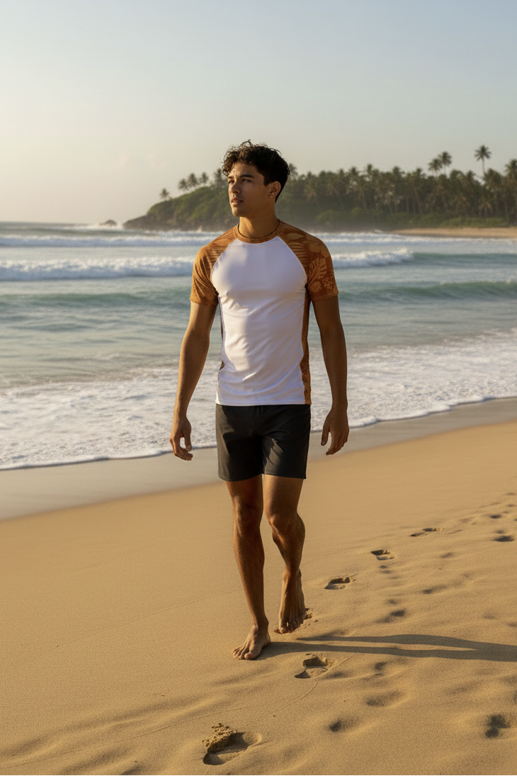 A man walking on a sandy beach wearing a Majestic Vision white and Clay Hibiscus rash guard with black boardshorts, with ocean waves and palm trees in the background.