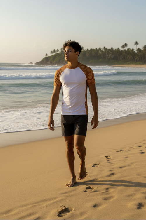 A man walking on a sandy beach wearing a Majestic Vision white and Clay Hibiscus rash guard with black boardshorts, with ocean waves and palm trees in the background.